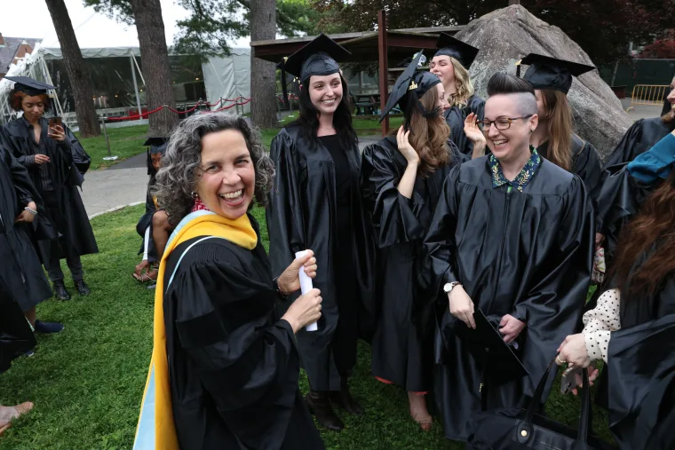 Group of graduates in caps and gowns laughing and talking outdoors, with one smiling at the camera.