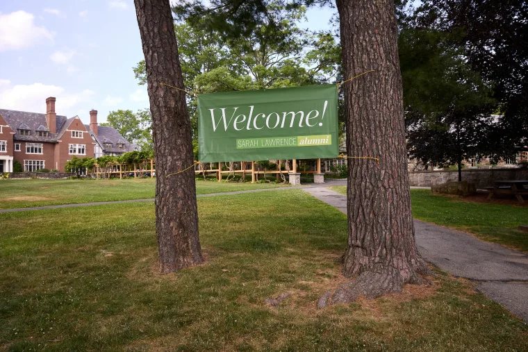 Banner reading "Welcome! Sarah Lawrence alumni" is hung between two trees on a grassy area near a historic brick building.