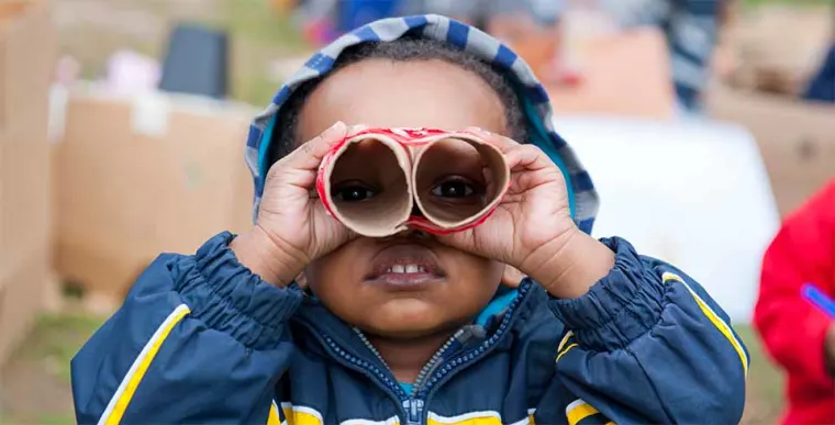 Child with a hooded jacket uses cardboard tubes as pretend binoculars, standing outside with blurred figures in the background.