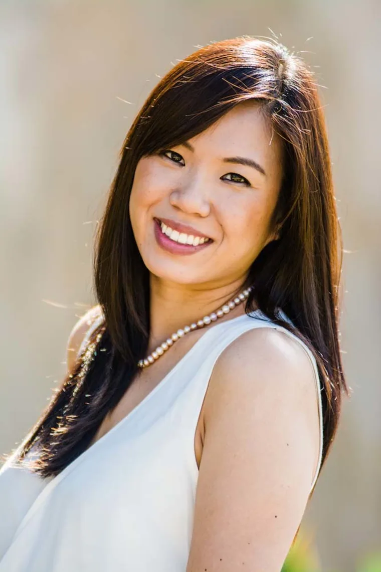 Person smiling, wearing a white top and pearl necklace, with long dark hair, standing outdoors with a blurred background.