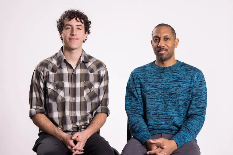 Two individuals sitting side by side, one in a plaid shirt and the other in a blue sweater, against a plain background.