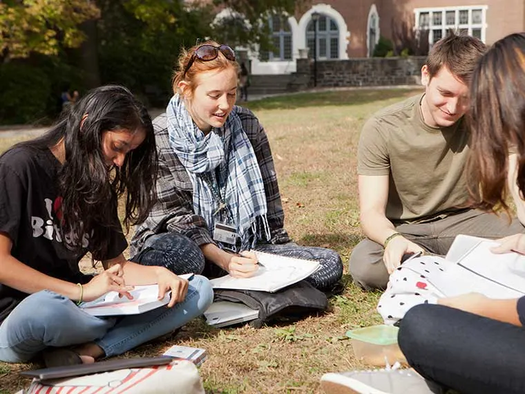 Group of people sitting on grass, drawing in notebooks, enjoying sunlight. Brick building with large windows in background.