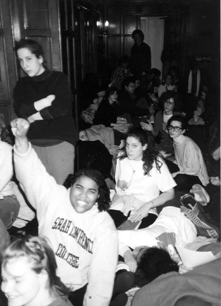 Group of people sitting closely together indoors, one person raising a fist and smiling, wearing a "Sarah Lawrence College" sweatshirt.