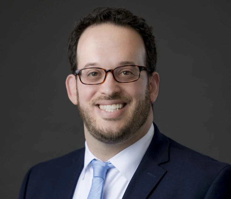Person with glasses, short hair, and beard, wearing a suit and tie, smiling against a dark background.