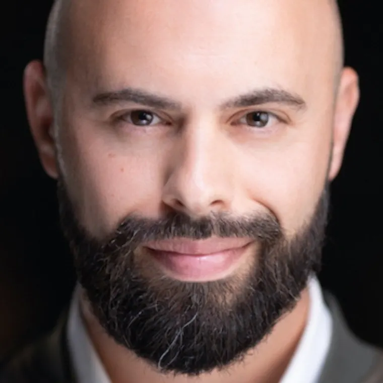 Person with a bald head and full beard, smiling softly, dressed in a collared shirt against a dark background.