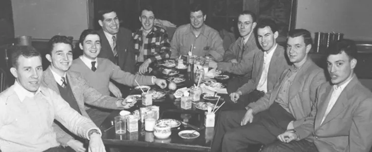 Group of ten people sitting around a dining table with drinks and plates. They are dressed in 1950s-style clothing.