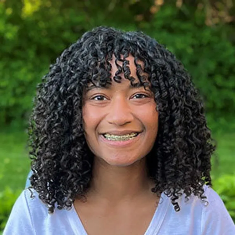 A person with curly hair and braces smiles outdoors with a green, leafy background.