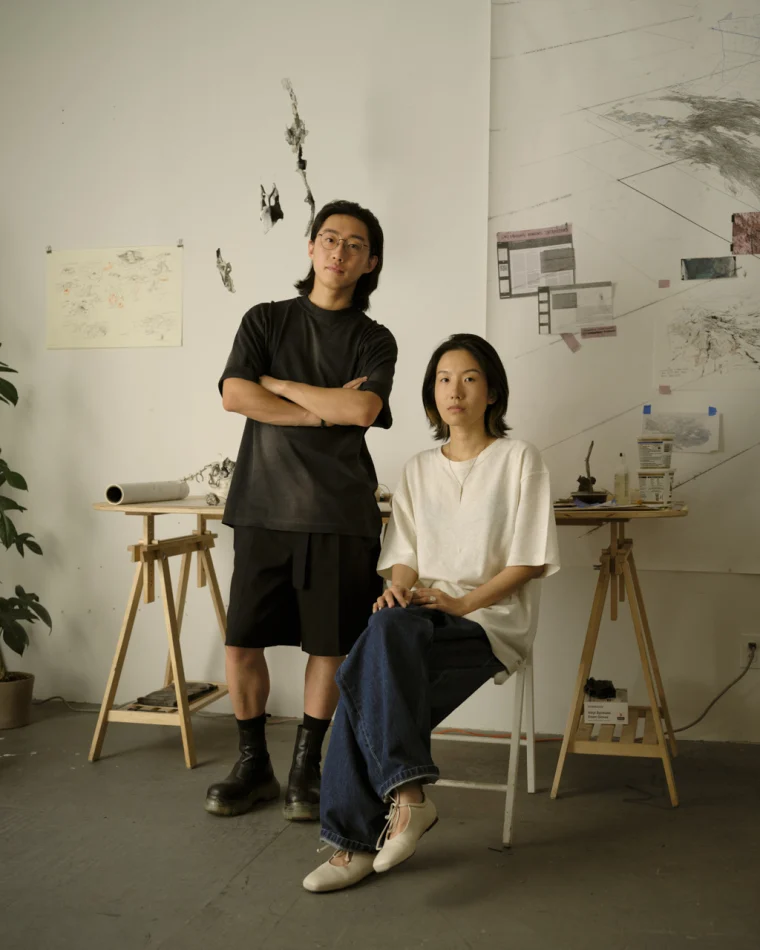 Two individuals in an art studio, one standing with arms crossed and the other seated. Artworks and tools are visible in the background.