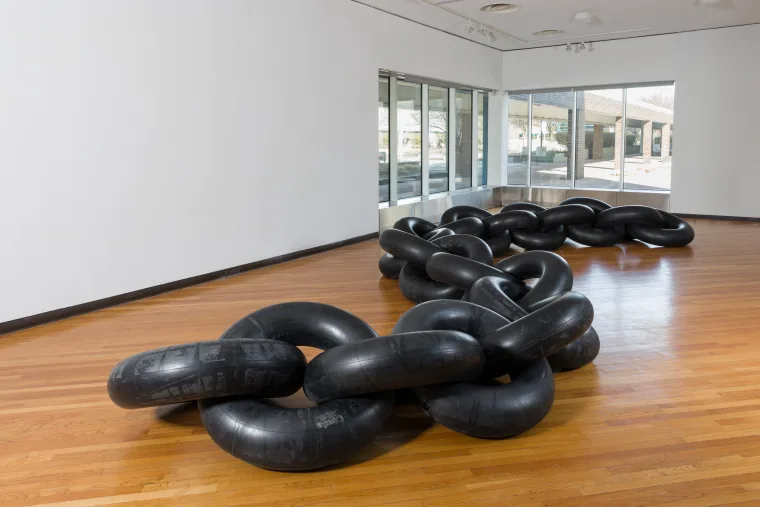 Art installation of large, black, inflated inner tubes linked together, displayed on a wooden floor in a bright gallery space.