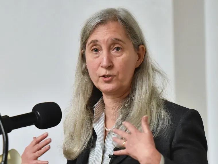 Person with long gray hair speaking at a microphone, wearing a suit jacket and gesturing with one hand.