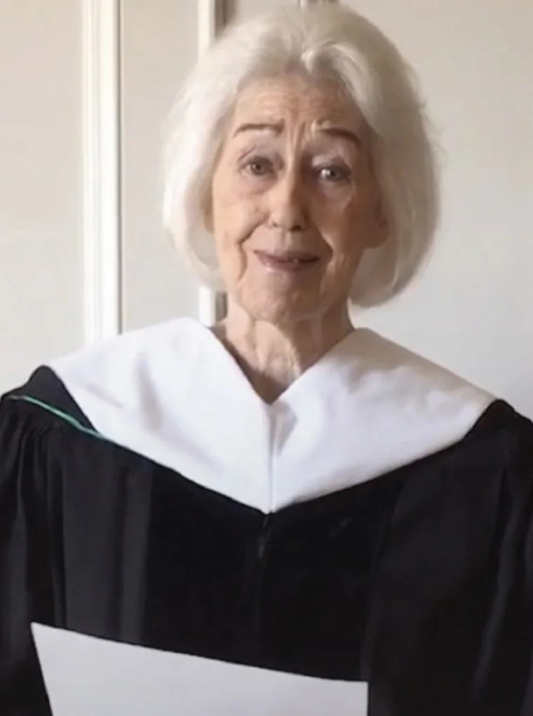 Person with white hair in graduation attire holding a paper, standing indoors against a light-colored wall.