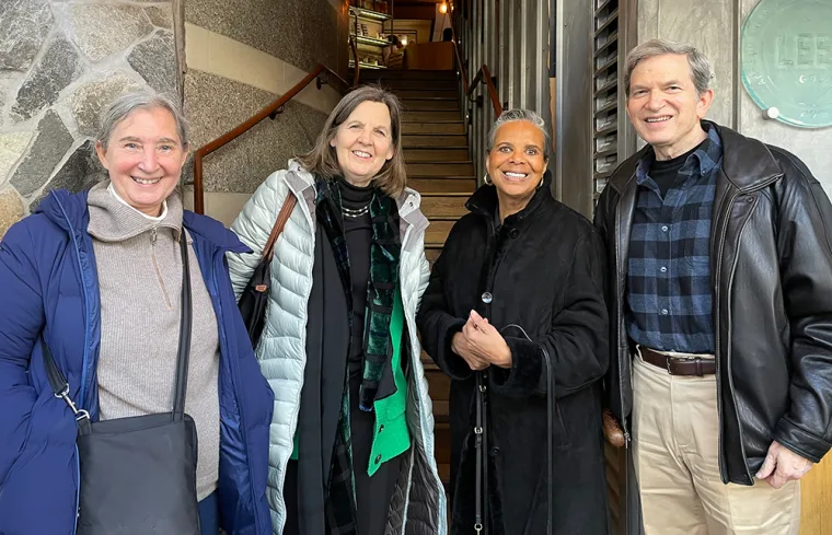 Four people standing together, smiling in front of a building entrance with stone walls and a staircase in the background.