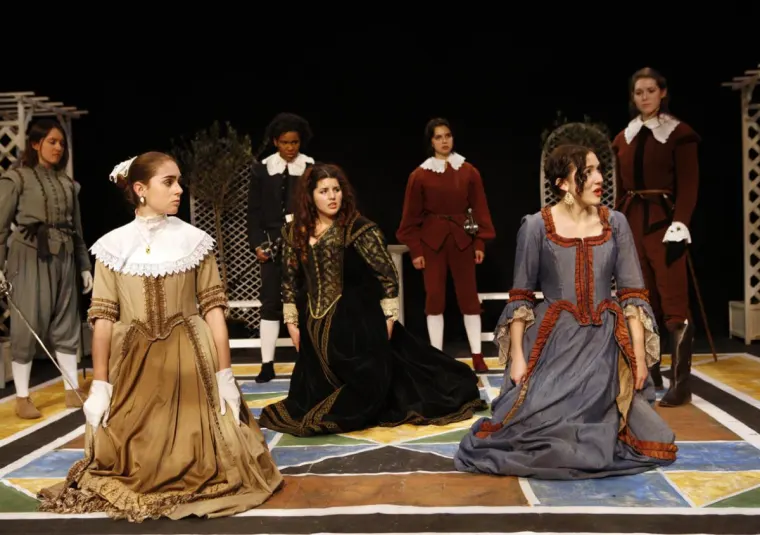 Seven people in period costumes stand and kneel on a colorful stage, with expressions of concern and focus. Backdrop includes lattice structures.