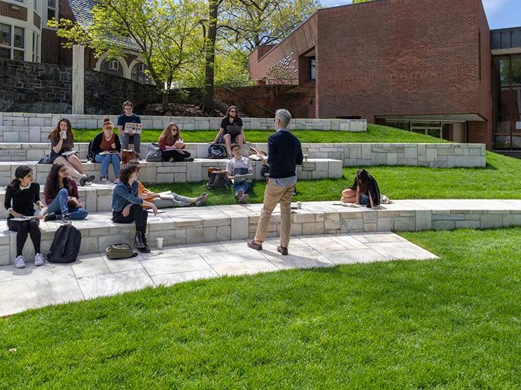 A group of people sit on tiered stone benches listening to a person standing in front of them. Green grass and a brick building are visible.