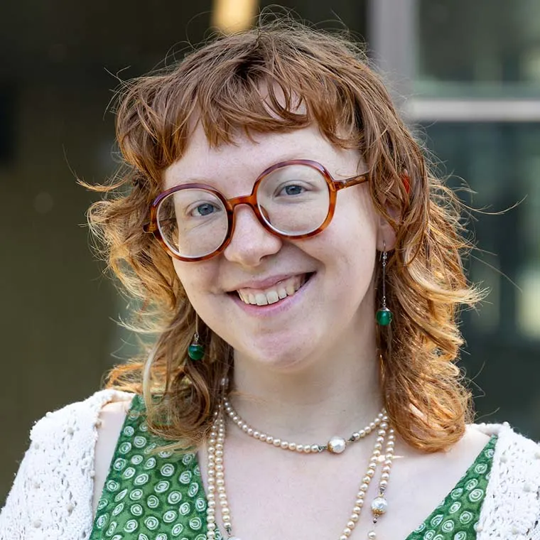 Smiling person with curly red hair, large round glasses, and wearing green clothing with pearl necklaces and beaded earrings outdoors.
