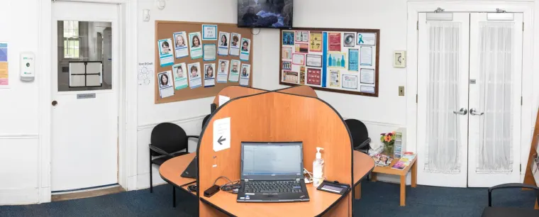 Office lobby with a workstation featuring a laptop, sanitizer, and bulletin boards covered with photos and notices. White doors are in the background.
