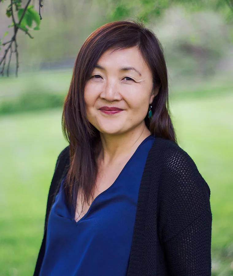 Person with long hair wearing a blue shirt and black cardigan, outside in a grassy area with blurred trees in the background.