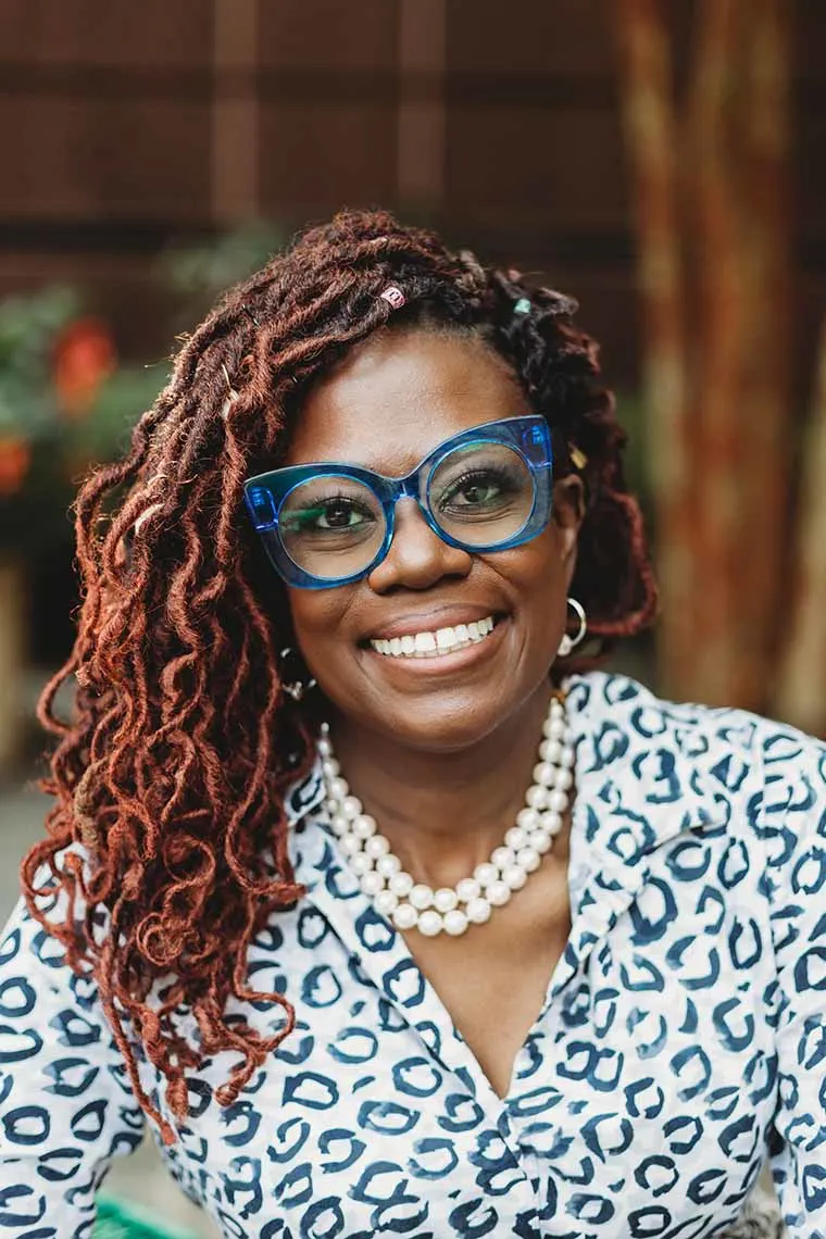 Person smiling, wearing large blue glasses, patterned shirt, and a pearl necklace, with red braided hair. Background is blurred and outdoors.