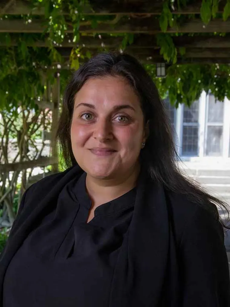 A person with dark hair smiles in an outdoor setting, surrounded by greenery and a wooden structure.
