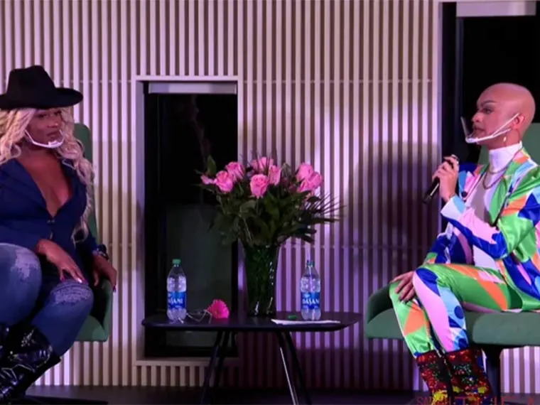 Two people sit on stage with a vase of pink roses between them. One wears a black hat, the other a colorful outfit, both with clear face shields.