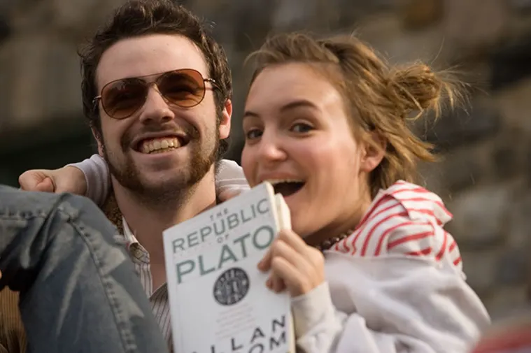 Two people smiling, one wearing sunglasses, the other holding a copy of "The Republic of Plato" in front of a blurred stone background.