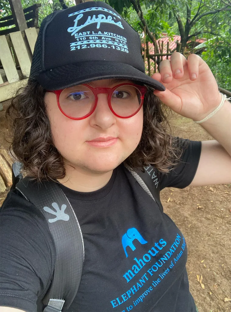 Person with curly hair, red glasses, black cap, and black shirt with an elephant graphic, outdoors holding the cap's brim.