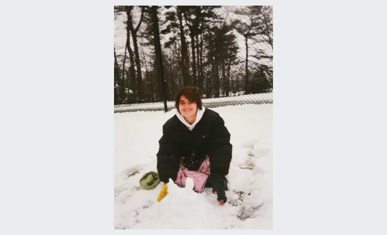 Person kneels in snow wearing black coat, surrounded by trees and a fence. A yellow glove and green item are nearby.