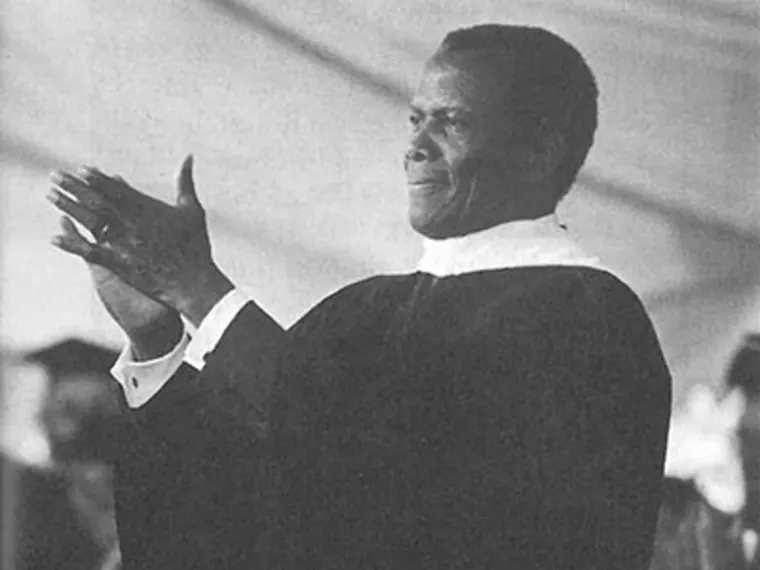 Person in academic gown clapping, with blurred figures in the background during what appears to be a formal event or ceremony.