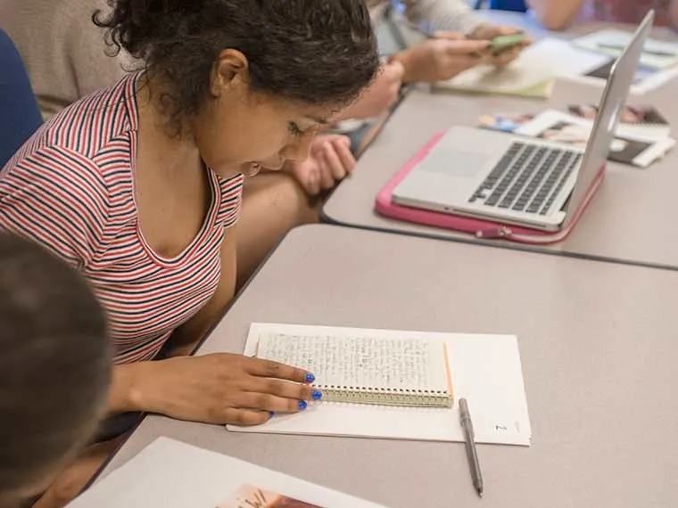 Person reading a notebook at a table with a laptop and other people nearby.