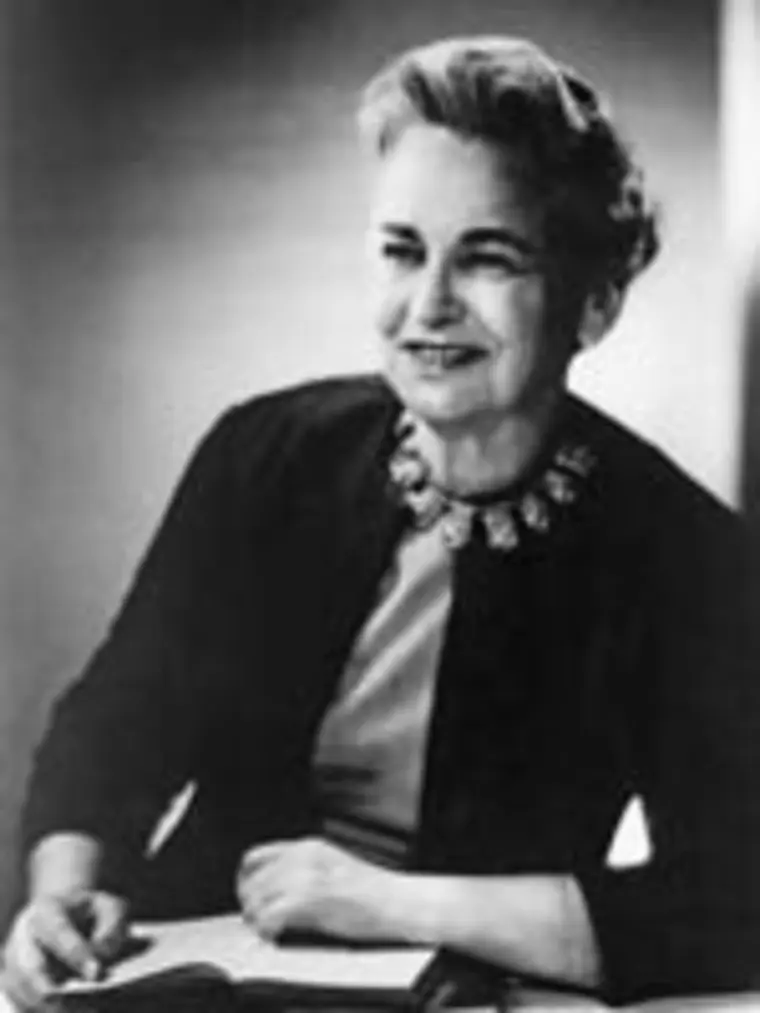 Person seated, smiling, wearing a dark jacket and floral necklace, holding a book. Black and white portrait.