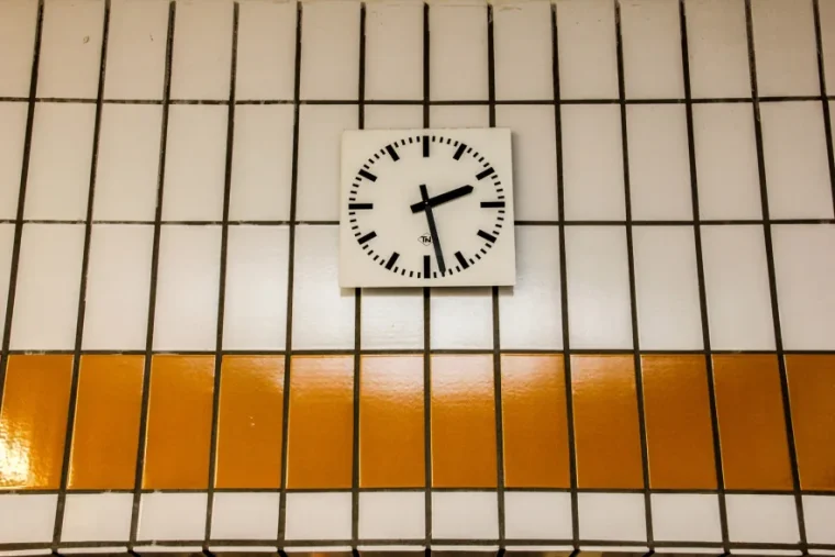 A wall clock with black hands and hour marks is mounted on a tiled wall with white and orange tiles.