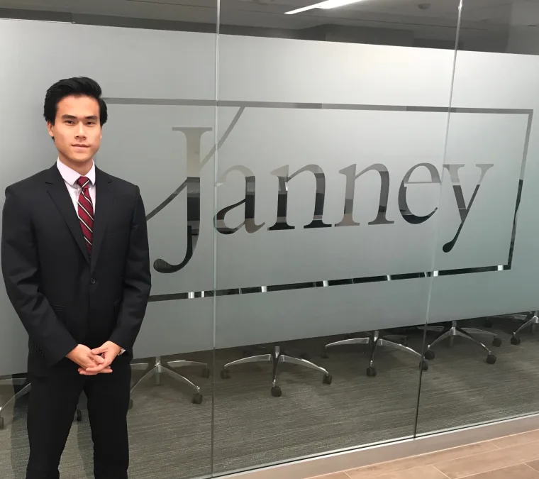 A person in a black suit and red tie stands in front of a glass wall with "Janney" written on it.
