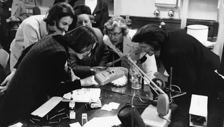 A group of people gather around a table, examining a handheld device, with scientific equipment and papers scattered around.