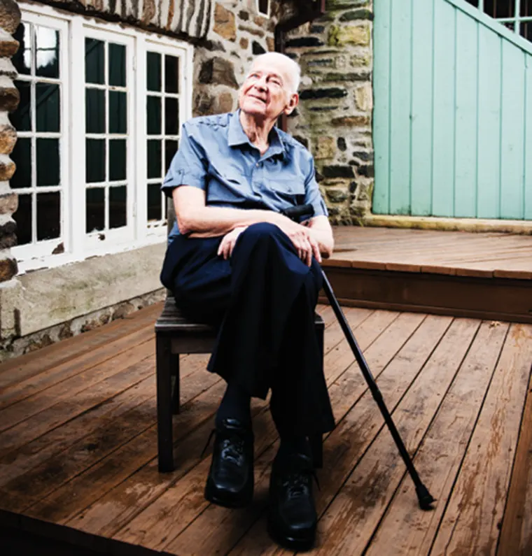 A person sitting on a wooden chair outside a rustic building, holding a walking stick and looking upwards.