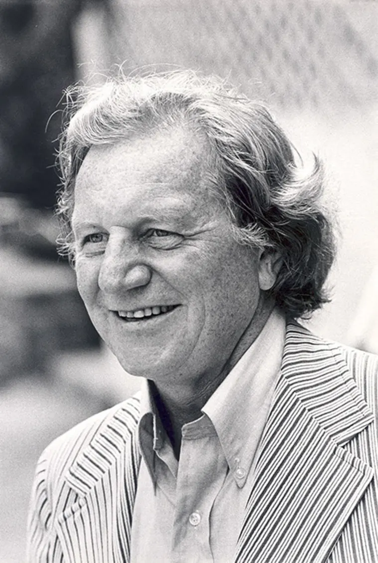 Smiling person with wavy hair in a striped blazer and collared shirt. Black and white photo with blurred background.