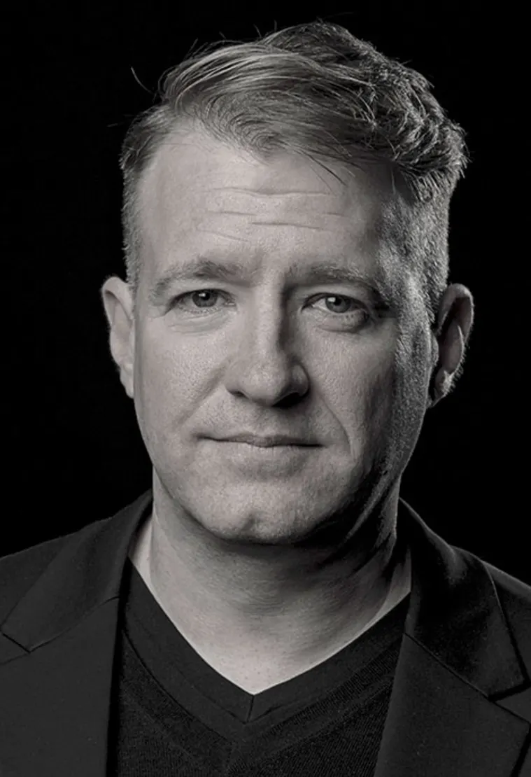 Black and white portrait of a person with short hair wearing a dark V-neck shirt and blazer, looking directly at the camera against a dark background.
