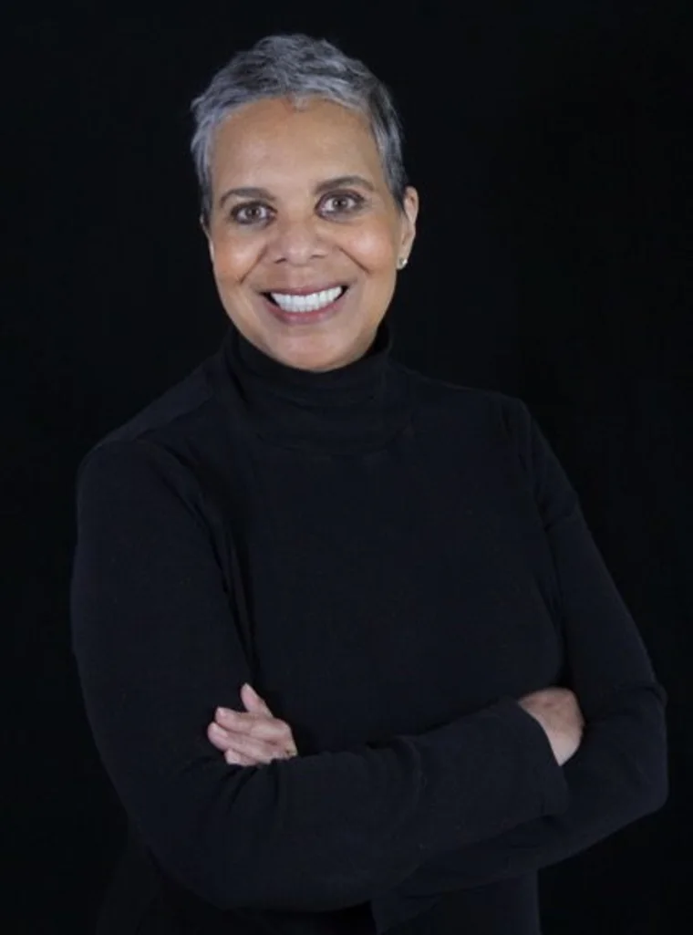 Person with short gray hair smiling, wearing a black turtleneck, against a black background, with arms crossed.