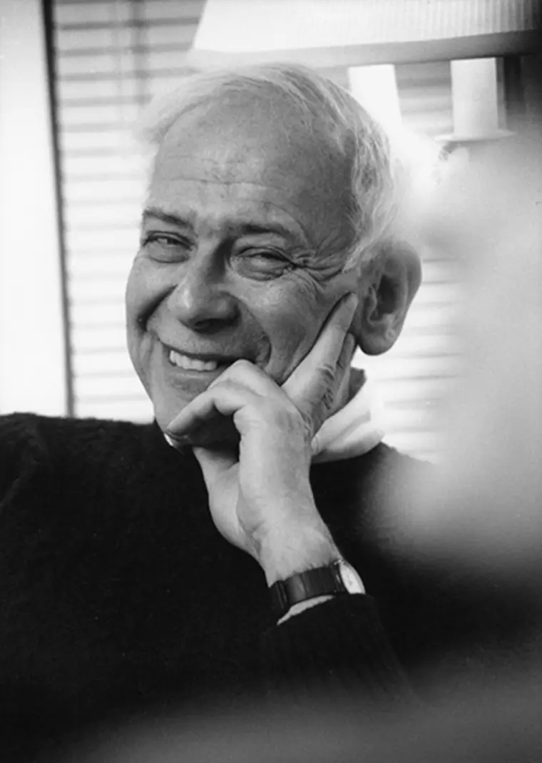 A smiling person with short, light hair rests their chin on their hand. Blinds and a lamp are visible in the softly focused background.