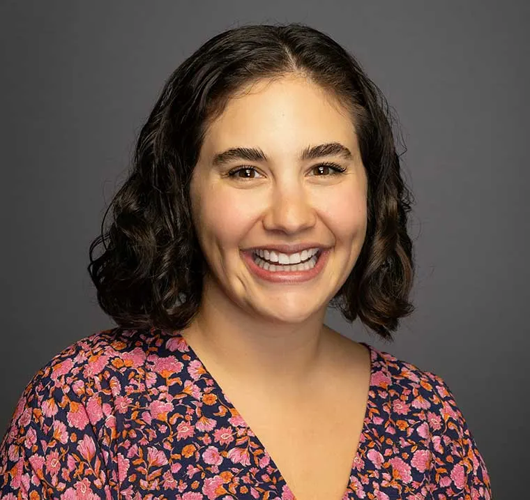 Person with curly hair smiling, wearing a floral-patterned top against a gray background.