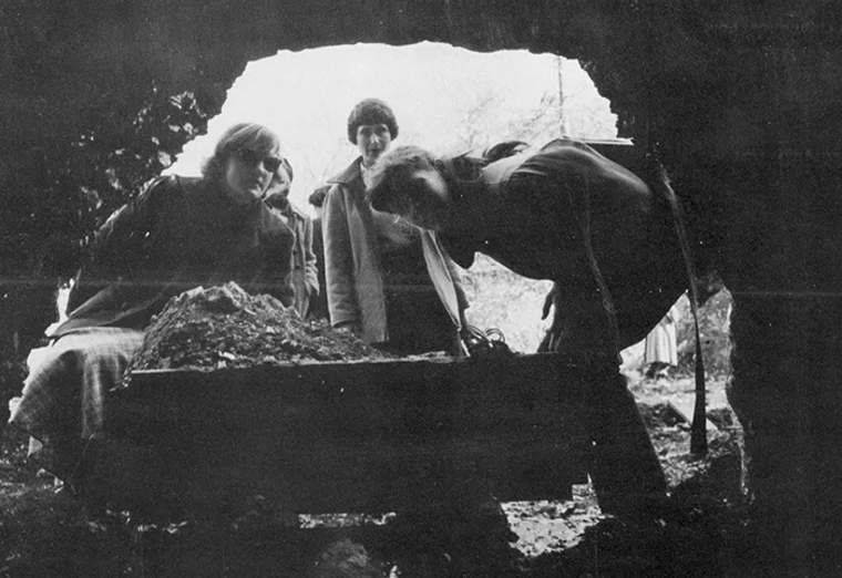 Three people peer into a cave from outside, examining rocks. The scene is captured in black and white.