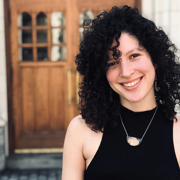Person with curly hair smiling in front of a wooden door, wearing a sleeveless black top and a pendant necklace.