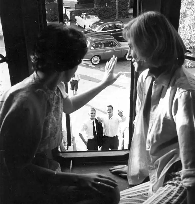 Two people on a windowsill look at and wave to three individuals outside on a street, surrounded by parked cars.