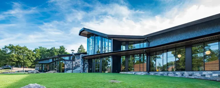 Modern building with large glass windows, sloped roof, and stone accents surrounded by green grass and trees under a blue sky.