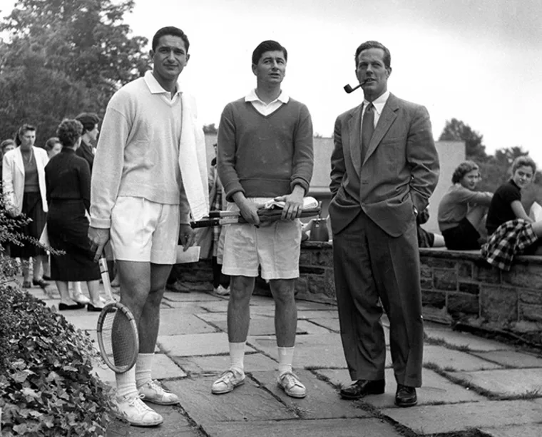 Three people stand outdoors, two in tennis attire holding rackets, one in a suit with a pipe. A group is gathered in the background.