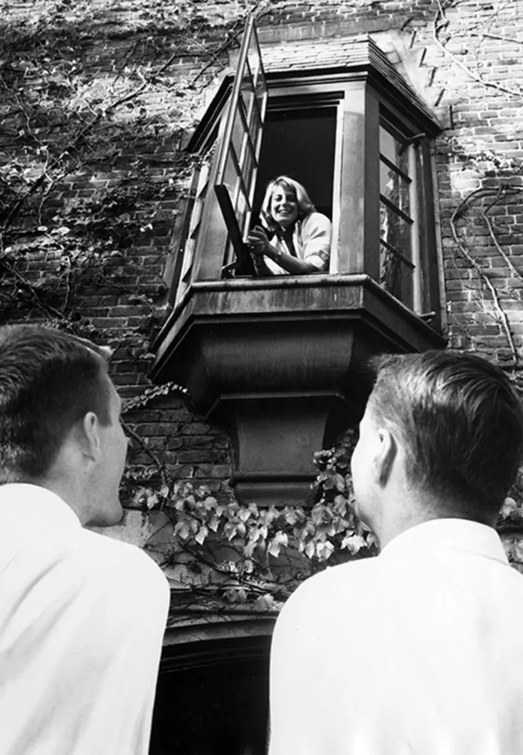 Person leaning out an open window, smiling at two people below, with ivy-covered brick wall in the background.