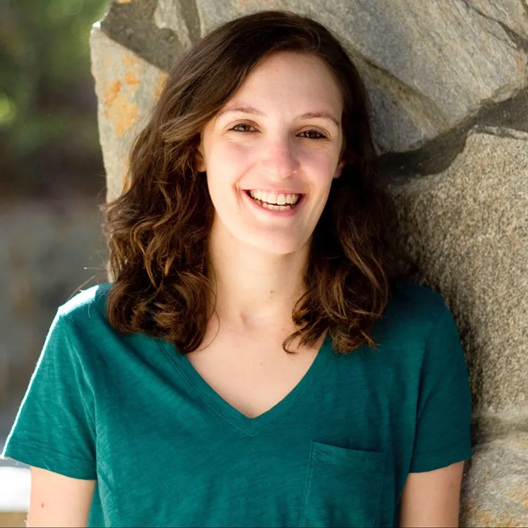 Person with wavy hair smiling, wearing a green shirt, stands against a stone wall in natural light.