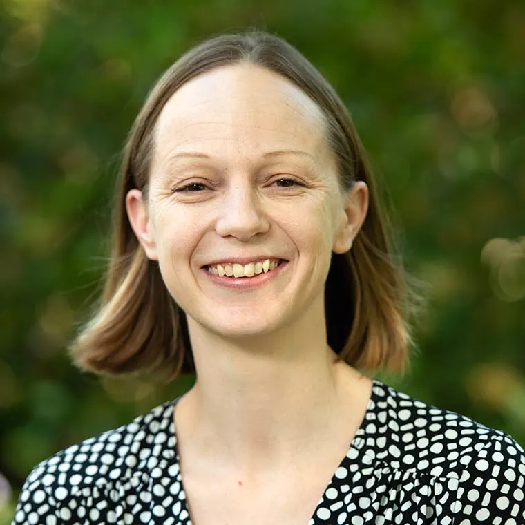 Smiling person with short hair, wearing a black and white polka dot shirt, against a blurred green background.