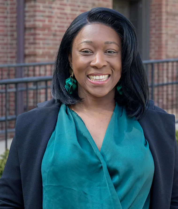 Smiling person wearing teal blouse and earrings, with dark hair, standing in front of a brick building and metal railing.