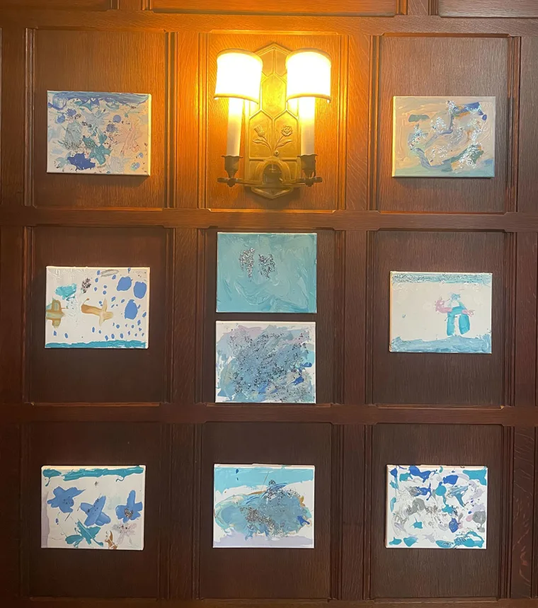 Nine abstract paintings with blue hues are arranged in a grid on a wooden wall, illuminated by a warm wall sconce.