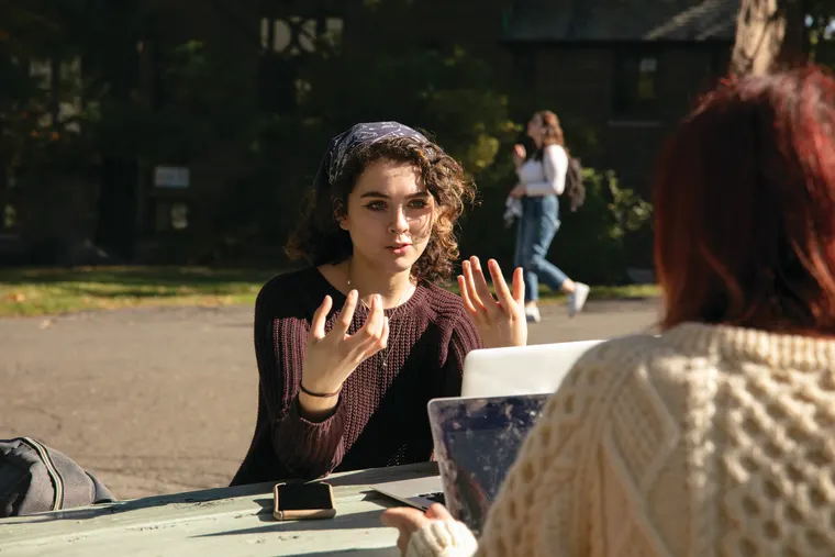 Two people sit at an outdoor table, engaged in conversation. One gestures while the other faces away, using a laptop.